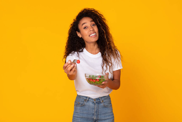 Улыбающаяся черная женщина с блюдом из овощного салата предлагает вилку для вас, рекомендуя здоровое питание с низким содержанием калорий для потери веса и здоровья, позируя на желтом фоне студии. Концепция питания - Фото, изображение