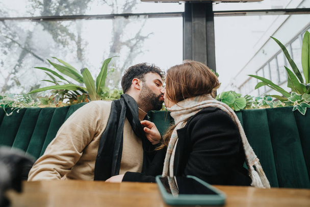 Ένα ερωτευμένο ζευγάρι μοιράζεται ένα φιλί σε ένα cafe της πόλης ανάμεσα σε πράσινα φυτά, συλλαμβάνοντας μια ρομαντική στιγμή σε ένα αστικό υπαίθριο περιβάλλον. - Φωτογραφία, εικόνα