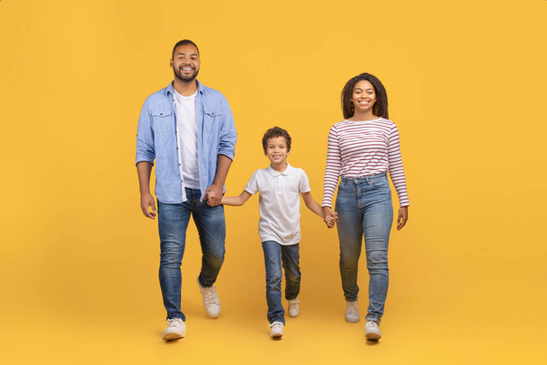 Zwarte familie van drie handen vasthouden en lopen over gele achtergrond, jonge Afrikaanse Amerikaanse ouders en tiener zoon in casual kleding belichamen ontspannen en gelukkige levensstijl, volledige lengte, kopieer ruimte - Foto, afbeelding