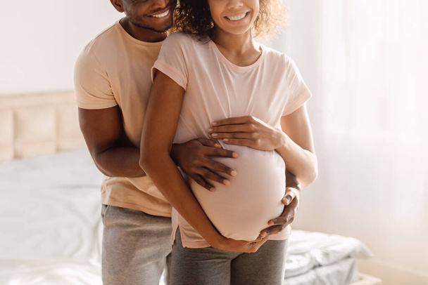 Концепция беременности. Обрезанный кадр молодого афроамериканца, ожидающего ребенка, муж обнимающий свою беременную жену и трогающий растущий живот в спальне в помещении - Фото, изображение