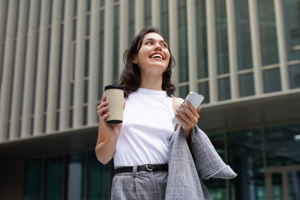 Europejska młoda kobieta biznesu szczęśliwie smsuje przez telefon, stoi przed urzędem miasta, sączy kawę podczas przerwy w pracy, łączy karierę z nowoczesnym miejskim stylem życia. Aplikacje i oferty mobilne - Zdjęcie, obraz