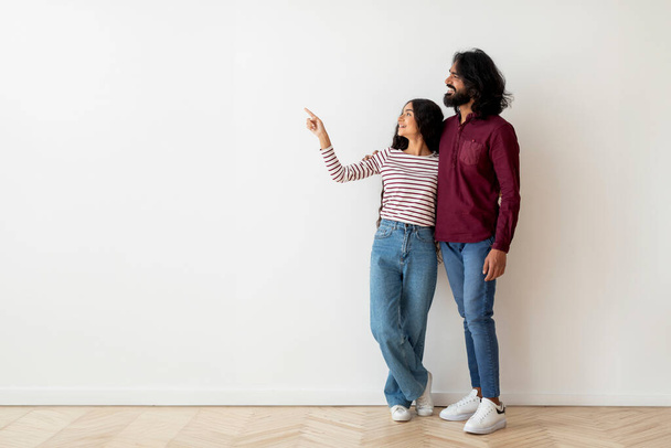 Loving millennial hindu couple happy man and woman wearing casual clothing embracing and pointing at copy space over white blank wall background in empty house, showing advertisement. Mortgage - Photo, Image
