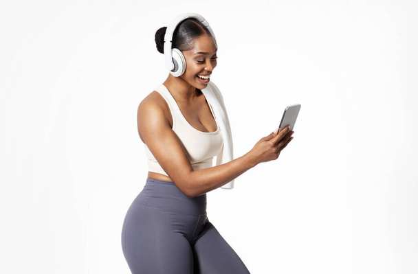 Mujer negra sonriente en ropa deportiva con auriculares escucha la canción en el teléfono, aislado en el fondo blanco, filmado en el estudio. Motivación del entrenamiento y música favorita para el entrenamiento de fitness - Foto, Imagen