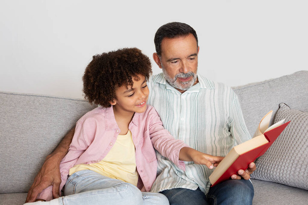 Латиноамериканец дедушка и внук глубоко погружены в книгу сказок, наслаждаясь совместным чтением, сидя с книгой на диване в доме, в удобной современной обстановке гостиной - Фото, изображение