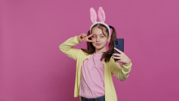 Confident happy young girl taking photos on smartphone webcam, feeling cute with her pigtails and bunny ears. Joyful smiling toddler takes pictures and fooling around in studio. Camera A. - Footage, Video