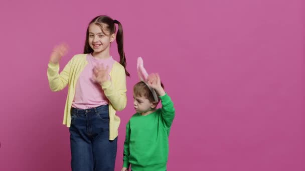 Joyful little kids waving in front of camera during easter holiday, smiling and wearing bunny ears. Brother and sister toddlers greeting someone in studio, adorable happy children. Camera A. - Footage, Video