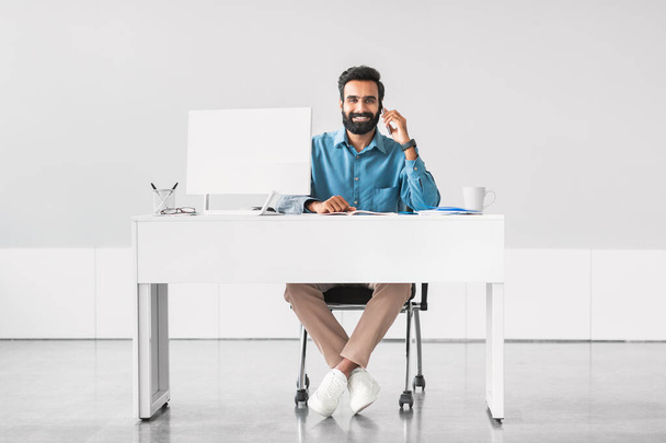 Brodatego hinduskiego biznesmena w niebieskiej koszuli, uśmiechniętego przez telefon, siedzącego wygodnie przy minimalistycznym biurku z komputerem w nowoczesnym biurze - Zdjęcie, obraz