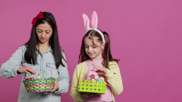 Joyful confident child and mother showing easter baskets on camera, decorating festive arrangements for spring holiday. Happy schoolgirl with bunny ears posing with her mom in studio. Camera A. - Footage, Video