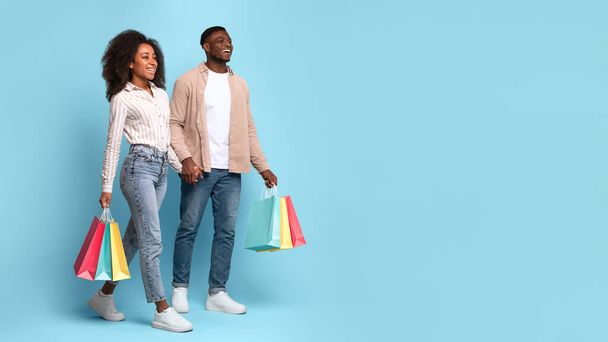 Fröhliches schwarzes Paar, lässig mit bunten Einkaufstüten spazierend, mit glücklichen Gesichtsausdrücken vorausblickend, vor blauem Hintergrund, Panorama mit freiem Raum - Foto, Bild