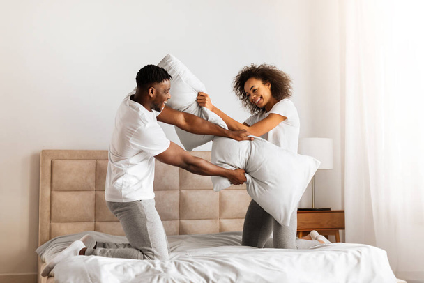 Wesoła Czarna Młoda Para Śmiejąca się i Flirtująca W Sypialni W Domu. Młody mąż i żona walczą razem z poduszkami dla zabawy w weekend. Szczęśliwego związku - Zdjęcie, obraz