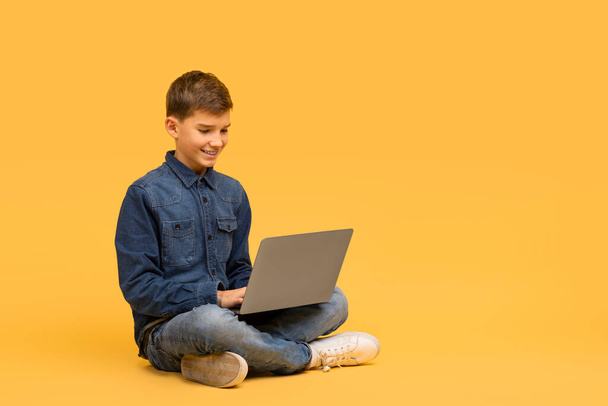 Усміхнений хлопчик-підліток сидить на перехресті ноги на підлозі і використовує ноутбук, щасливий чоловік з комп'ютерним навчанням онлайн, позує на жовтому студійному фоні, насолоджуючись сучасними технологіями, копіюючи простір - Фото, зображення