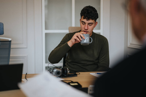 Un giovane uomo sorseggia un caffè durante una pausa di lavoro, mostrando un momento di relax in mezzo a una giornata di lavoro in ufficio. - Foto, immagini