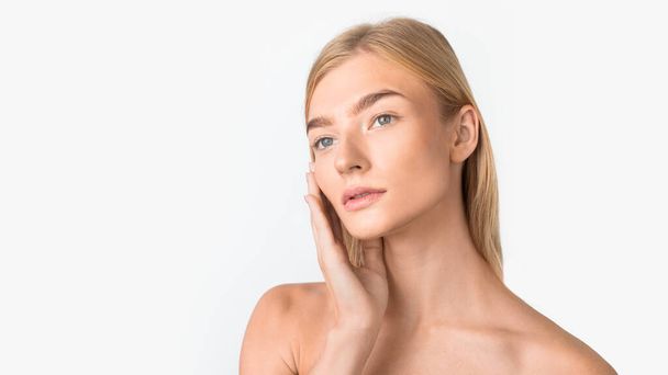 Facial Spa concept. Gorgeous blonde lady touching her soft skin on cheek, enjoying result of beauty treatment, looking aside, standing shirtless near free space on white studio background. Panorama - Photo, Image