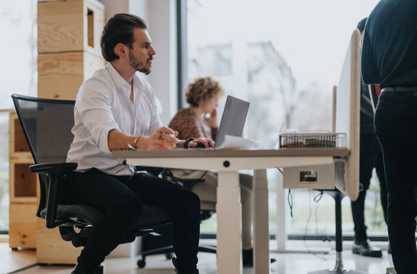 Een professionele man concentreert zich op het werk op zijn laptop in een levendige kantooromgeving, met collega 's die zich bezighouden met taken om hem heen. - Foto, afbeelding