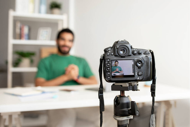 Homme Moyen-Orient dans une chemise verte assis à un bureau avec un sourire confiant, capturé à travers un écran de caméras professionnelles en bref, dans une configuration de bureau à domicile pour l'enregistrement vidéo - Photo, image