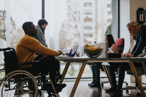 Inclusieve arbeidscultuur afgebeeld met multi-etnisch team, waaronder een persoon met een handicap die deelneemt aan een gezamenlijke werksessie in een helder kantoor. - Foto, afbeelding