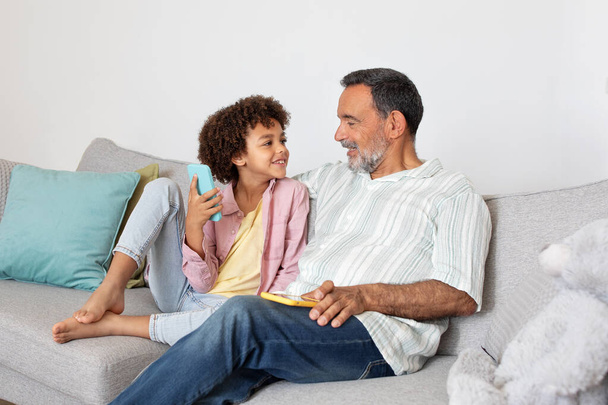 Latein Opa und Enkelkind glücklich mit Smartphone-Anwendung, sitzen auf dem Sofa und surfen im Internet mit Handy-Gerät drinnen. Moderne Großelternschaft, Familie und Gadgets - Foto, Bild