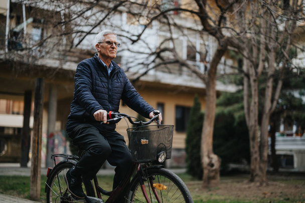 Ενεργός ηλικιωμένος κύριος ποδηλασία σε εξωτερικούς χώρους, επιδεικνύοντας έναν υγιεινό τρόπο ζωής και δραστηριότητες αναψυχής για ηλικιωμένους σε ένα αστικό πάρκο ρύθμιση. - Φωτογραφία, εικόνα