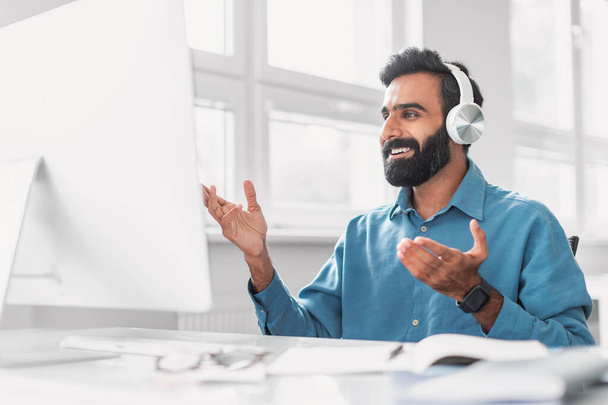 Hombre barbudo indio comprometido con camisa azul con auriculares gesticulantes, completamente inmerso en una animada conversación virtual en un entorno de oficina brillante - Foto, Imagen