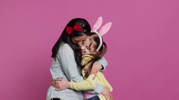 Lovely schoolgirl with bunny ears an her mom waving on camera, having fun and laughing against pink background. Cheerful mother and her daughter embracing and kissing each other. Camera A. - Footage, Video