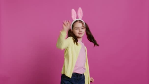 Chica alegre y confiada mostrando movimientos de baile en el estudio, sintiéndose alegre y positiva sobre la festividad navideña de Pascua. Niño talentoso bailando y usando orejas de conejo. Cámara B. - Metraje, vídeo