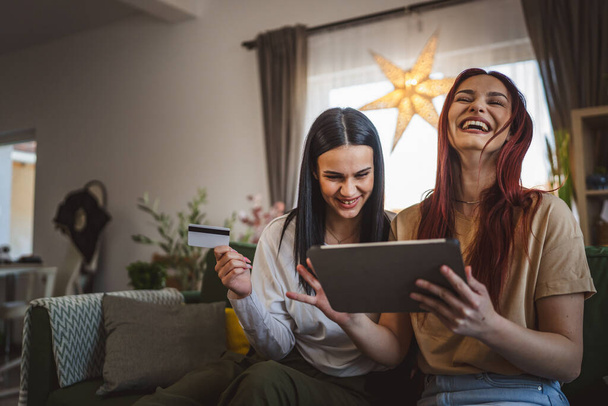 δύο νεαρές γυναίκες αδελφές ή φίλοι κάθονται στο σπίτι χρήση πιστωτικής κάρτας για online ψώνια περιηγηθείτε στο διαδίκτυο καταστήματα σε ψηφιακή ταμπλέτα αγορά πράγματα έκπτωση πώληση πραγματικών ανθρώπων αντίγραφο χώρο - Φωτογραφία, εικόνα