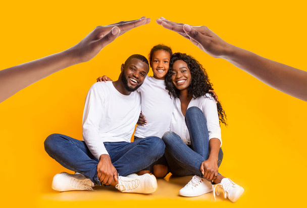 A radiant african american family of three, dressed in white tops and denim, sits closely, framed by hands forming a house shape above them on a vivid yellow background. Mortgage, real estate - Photo, Image
