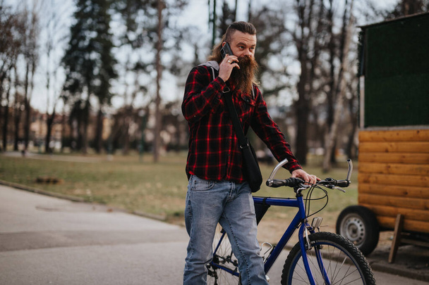 Κομψό αρσενικό επιχειρηματία χρησιμοποιώντας έξυπνο τηλέφωνο, ενώ στέκεται με το ποδήλατό του σε ένα αστικό πάρκο, επιδεικνύοντας σύγχρονη απομακρυσμένη εργασία. - Φωτογραφία, εικόνα