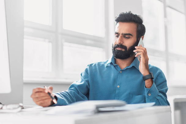 Uważny indyjski biznesmen w niebieskiej koszuli na poważnym telefonie, długopis w ręku, starannie przeglądając dokumenty w dobrze zorganizowanej przestrzeni biurowej - Zdjęcie, obraz