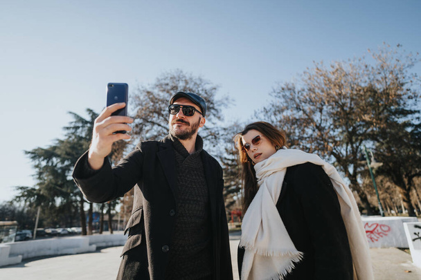 Dos jóvenes profesionales capturan un momento junto con una selfie. Vestidos con elegancia, disfrutan de un descanso en un soleado día de invierno en un entorno urbano. - Foto, imagen