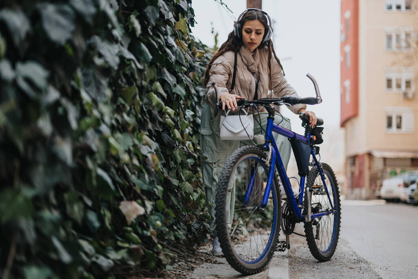 Skupiona młoda kobieta ze słuchawkami robi sobie przerwę od jazdy na rowerze w mieście, stojąc obok swojego niebieskiego roweru górskiego. - Zdjęcie, obraz