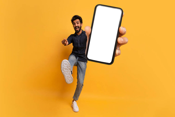Чудова онлайн угода. Веселий молодий індійський чоловік вказує на камеру і показує смартфон з порожнім екраном, жовтий студійний фон, повнометражний знімок, макет, простір для копіювання - Фото, зображення