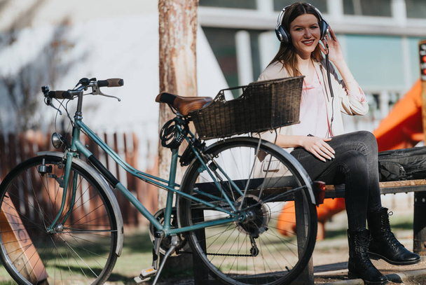 Una joven feliz se está relajando en un banco con sus auriculares, sonriendo mientras escucha música, bicicleta vintage estacionada en primer plano. - Foto, Imagen