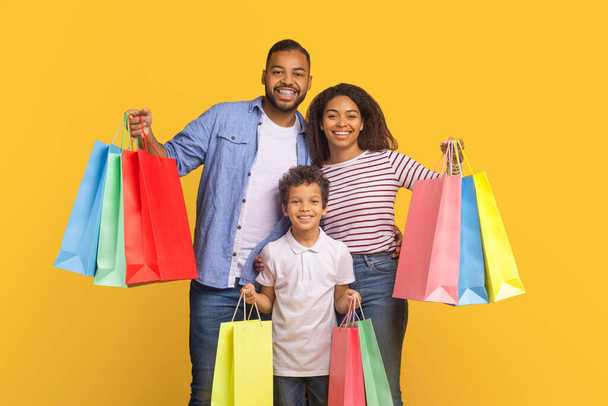 Familia negra de tres personas de pie con coloridas bolsas de compras en las manos, padres afroamericanos e hijo preadolescente sonriendo y disfrutando del día de compras, posando sobre un fondo amarillo brillante - Foto, imagen