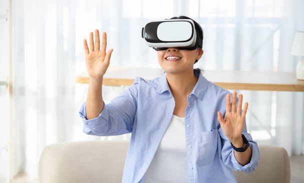 Gioioso asiatico giovane donna indossare moderno VR occhiali mentre seduto su divano in soggiorno, toccando spazio vuoto, cinese signora esperienza virtuale sessione di realtà a casa - Foto, immagini