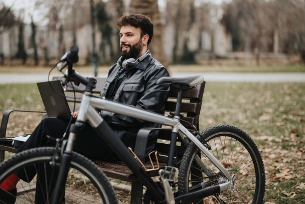 Κομψός επιχειρηματίας απολαμβάνει την απομακρυσμένη εργασία σε ένα πάρκο με το ποδήλατό του δίπλα του, χαρακτηριστικό παράδειγμα ενός ευέλικτου σύγχρονου τρόπου ζωής. - Φωτογραφία, εικόνα