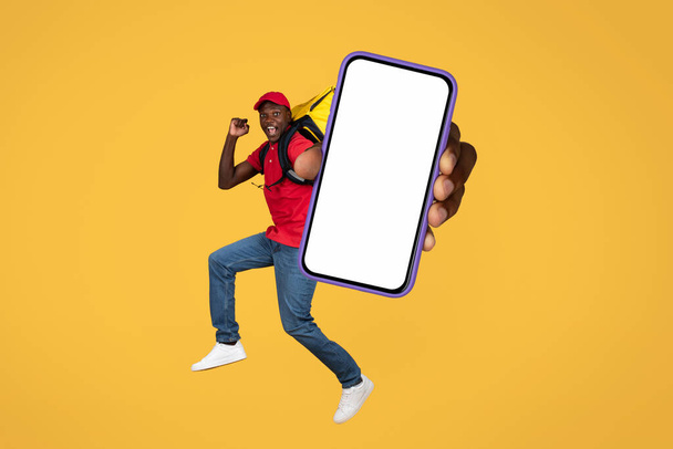 Ευτυχισμένος νεαρός Αφροαμερικάνος ντελιβεράς με στολή και σακίδιο να πηδάει στον αέρα, δείχνοντας μεγάλο τηλέφωνο με λευκή οθόνη, απομονωμένος σε πορτοκαλί φόντο στούντιο. Υπηρεσία παράδοσης παραγγελιών, εφαρμογή για κινητά - Φωτογραφία, εικόνα