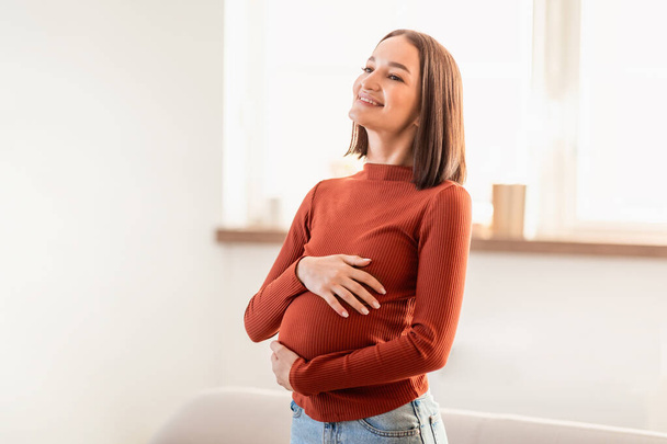Senhora expectante relaxado em pé em sua sala de estar em casa, sorri como ela sente seu movimento bebê tocando barriga, estimando momentos especiais de gravidez. Mulher grávida desfrutando de expectativa - Foto, Imagem