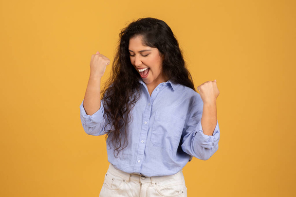 Exuberante mujer joven árabe alegre con el pelo rizado largo, con una blusa azul, celebra con los puños bombeados y los ojos cerrados, expresando alegría sobre un fondo amarillo, estudio - Foto, imagen