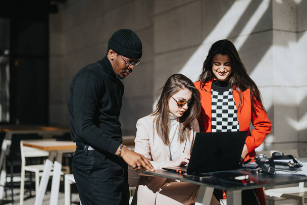 Trois jeunes gens multiraciaux collaborent à l'aide d'un ordinateur portable à une table de café extérieure ensoleillée, mettant en valeur le travail d'équipe et la créativité. - Photo, image