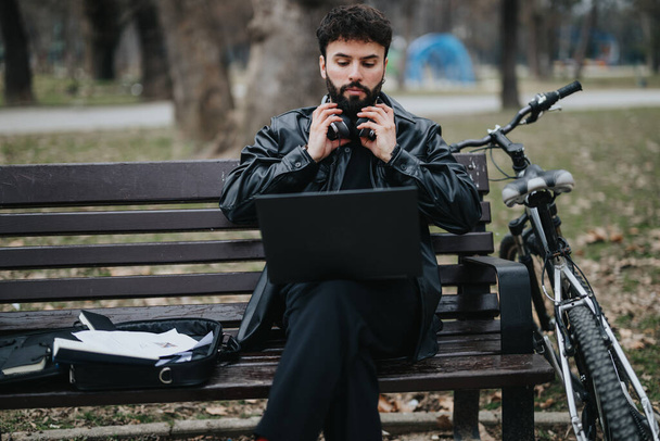 Empreendedor de negócios masculino moderno trabalhando ao ar livre com fones de ouvido e um laptop, sentado em um banco de parque ao lado de uma bicicleta. - Foto, Imagem