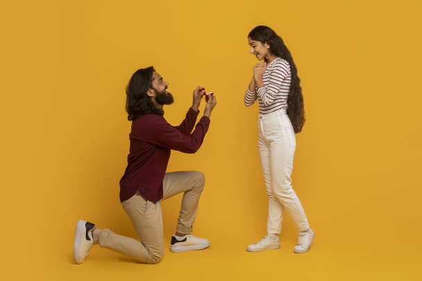 Liebevoll aussehender Indan-Mann macht seiner ziemlich aufgeregten Freundin einen Heiratsantrag, steht auf einem Knie und hält eine Schachtel mit Ring, gelbem Hintergrund. Verlobung, Ehe, Liebe - Foto, Bild
