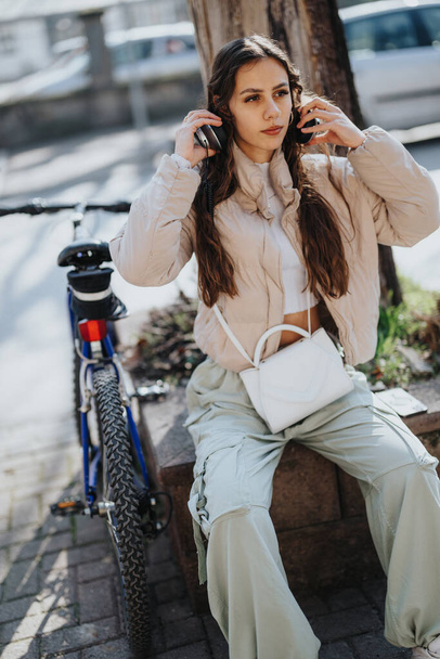 Stylish urban woman relaxing with her music, sitting by her bike on a sunny day in the city, capturing a moment of leisure. - Photo, Image