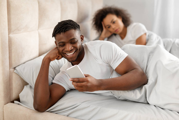 Phubbing. Niezadowolona Afroamerykanka patrząca na chłopaka piszącego przez telefon w łóżku, ignorującego ją w sypialni w domu. Zazdrosna pani podejrzewająca romans. Skupienie selektywne - Zdjęcie, obraz
