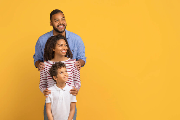 Χαρούμενη νεανική μαύρη οικογένεια των τριών που στέκεται στη σειρά και κοιτάζοντας πέρα από το χώρο των αντιγράφων, χαρούμενους γονείς και έφηβος γιος απόσπαση πάνω από κίτρινο φόντο στούντιο, απολαμβάνοντας νέα προσφορά ή Promo, Πανόραμα - Φωτογραφία, εικόνα
