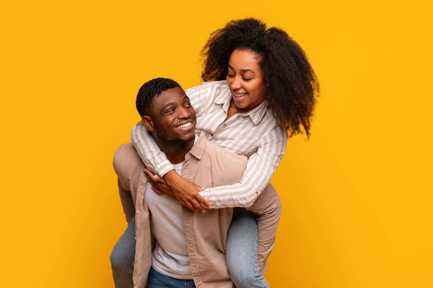 幸せなアフリカ系アメリカ人男性は,女性にピギーバックに乗って,陽気な黄色の背景に対して一緒に笑いと遊び心のある瞬間を楽しんでいます - 写真・画像