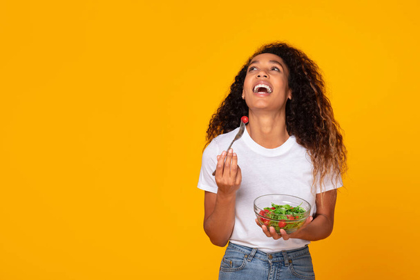 Schwarze junge Dame posiert mit einer gesunden grünen Salatmahlzeit, hält Schüssel und Gabel in der Hand und genießt lachend ihr Abendessen vor gelber Studiokulisse. Gewichtsverlust und Schlankheitswahn. Freiraum - Foto, Bild