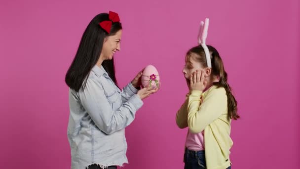 Urocza matka zaskakująca swoją córkę z uroczym różowym jajkiem na święto wielkanocne, śmiejąc się razem na różowym tle. Radosna dziewczyna jest zdumiona prezentem od mamy. Kamera B. - Materiał filmowy, wideo