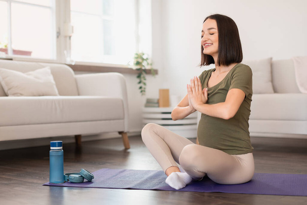 Embarazada practica yoga y meditación, sentada en pose de loto con las manos cerradas en el interior, ejercitándose y relajándose en casa. Relajación y bienestar durante el embarazo - Foto, imagen
