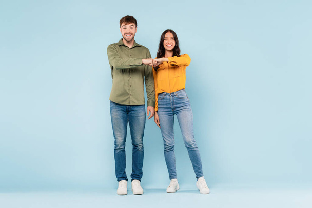 Lachende jonge man en vrouw in casual kleding vuist stoten in vriendelijke groet of overeenkomst, staan tegen zacht blauwe achtergrond. - Foto, afbeelding